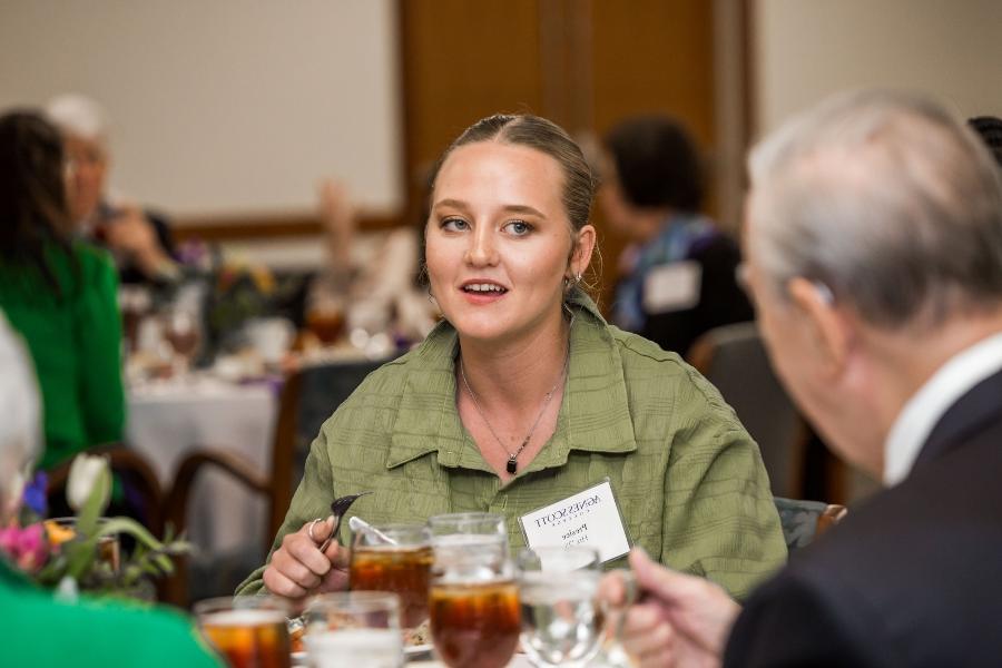 A young woman with blonde hair seated at a dining table, engaged in conversation. She is wearing a green button-up shirt and a name tag for Agnes Scott College. There are plates and glasses of iced tea on the table in front of her, and other attendees are visible in the background.